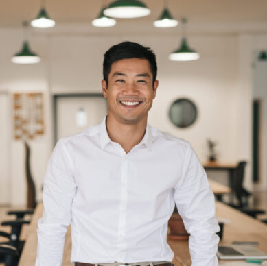 Smiling young Asian businessman leaning against a table while working alone in a large modern office