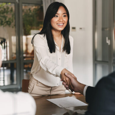 Business, career and placement concept - image from back of two employers sitting in office and shaking hand of young asian woman after successful negotiations or interview